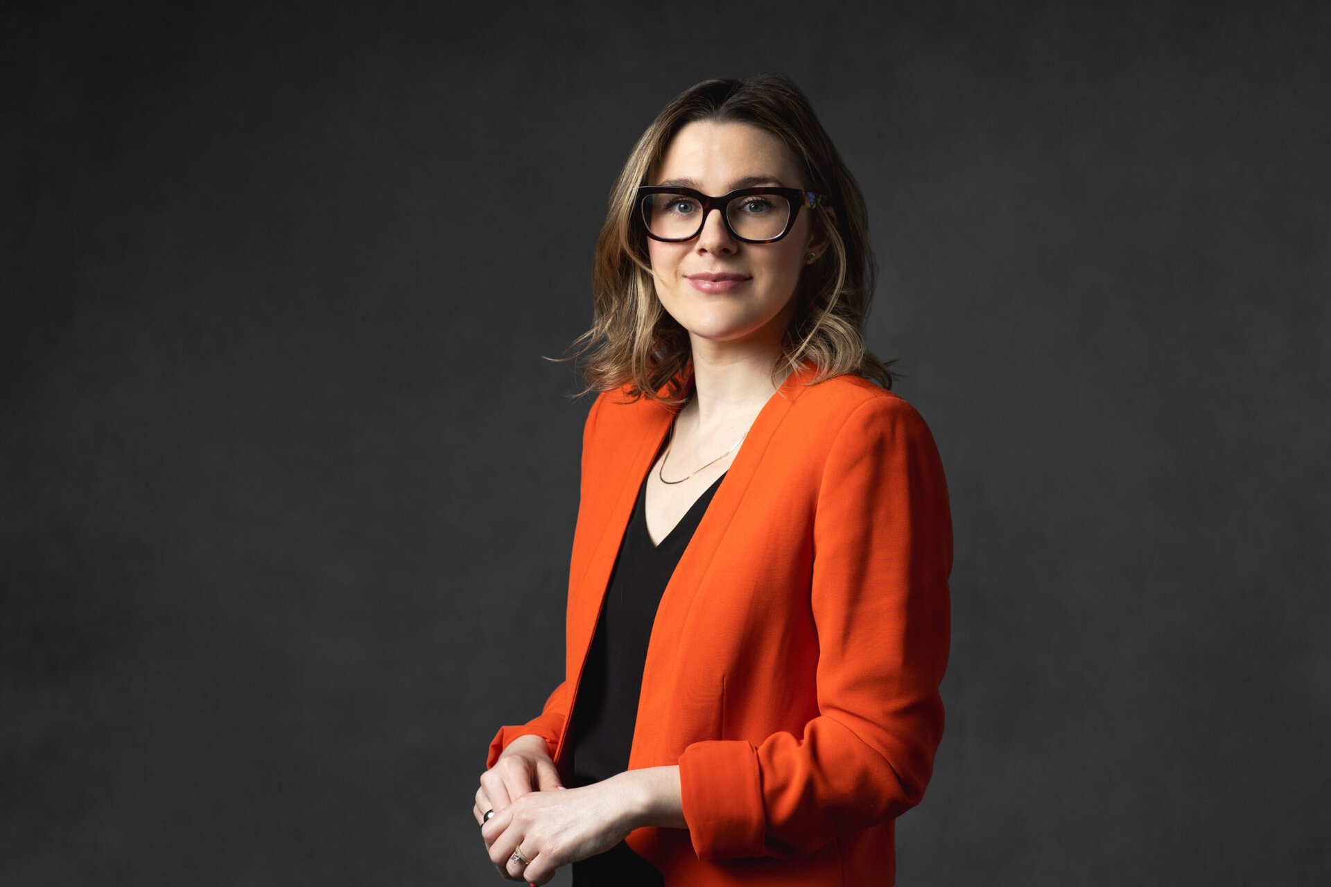 Professional woman in orange blazer and glasses against dark background.